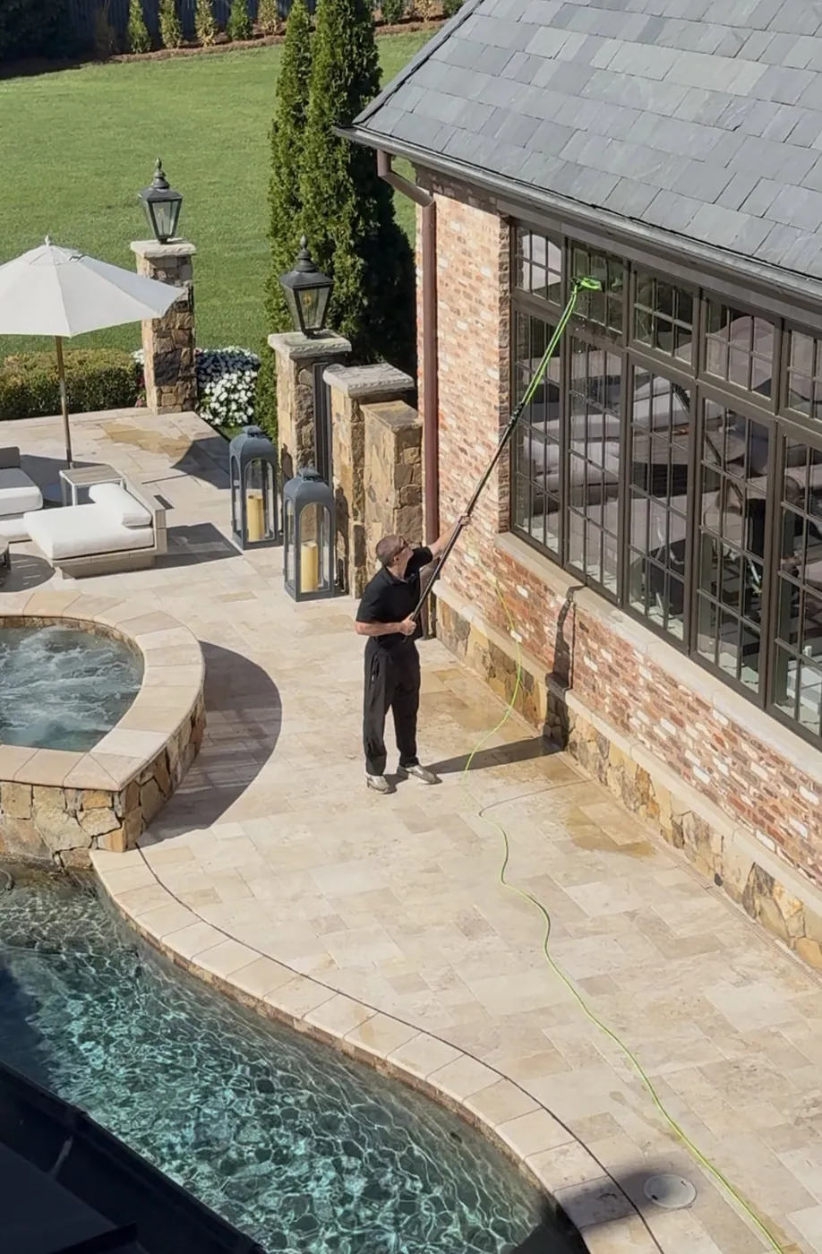 Technician using a waterfed pole to clean the tall windows of a brick pool house beside a swimming pool.